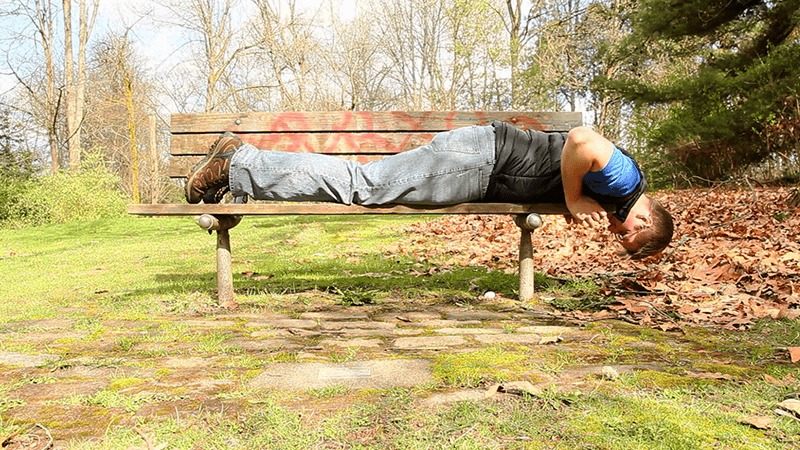 A geocacher lies across a park bench and reaches underneath it to try and find a geocache.